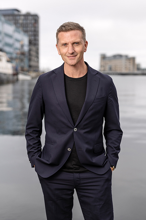 En man med kostym står vid vattnet med händerna i fickorna